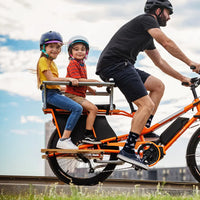 Vélo cargo longtail électrique Yuba Kombi Orange tout équipé en situation avec des enfants et un père